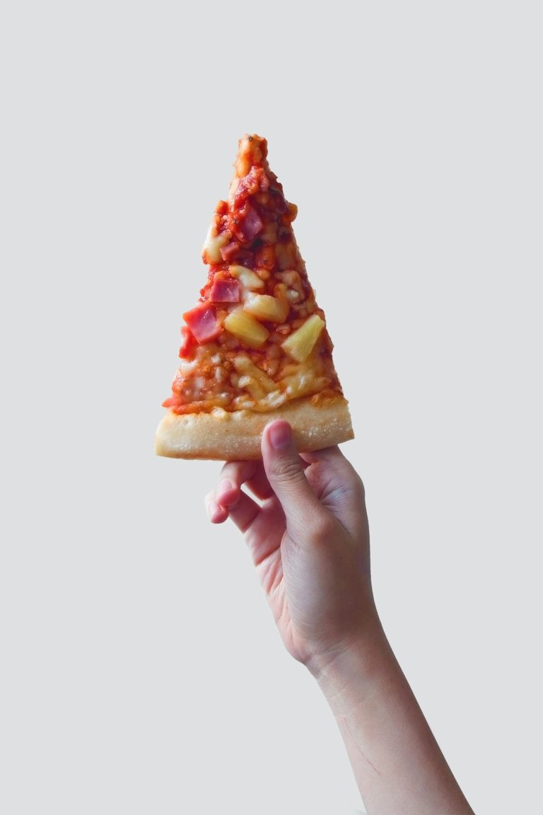Hand holding up a slice of pineapple and ham pizza against a white background